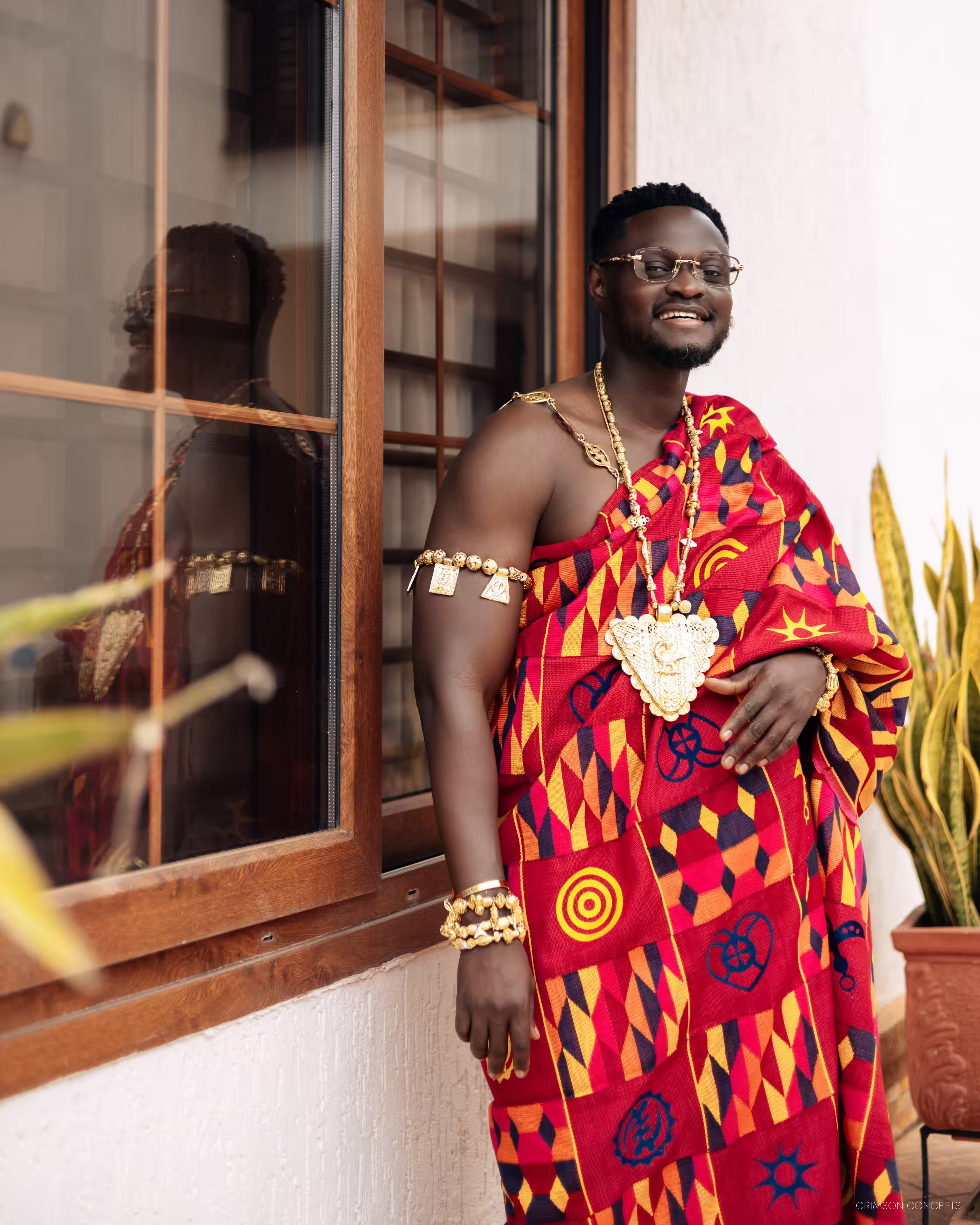 Qojoh smiling beside a window in his red and gold kente cloth